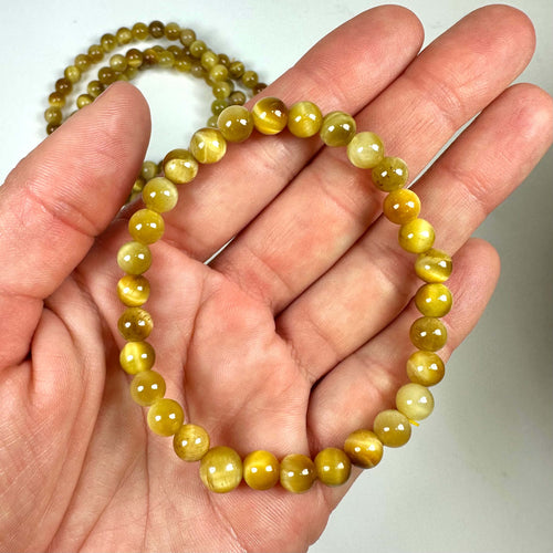En hånd holder et Gul Tigerøje armbånd med runde perler, mens et andet armbånd ses i baggrunden.