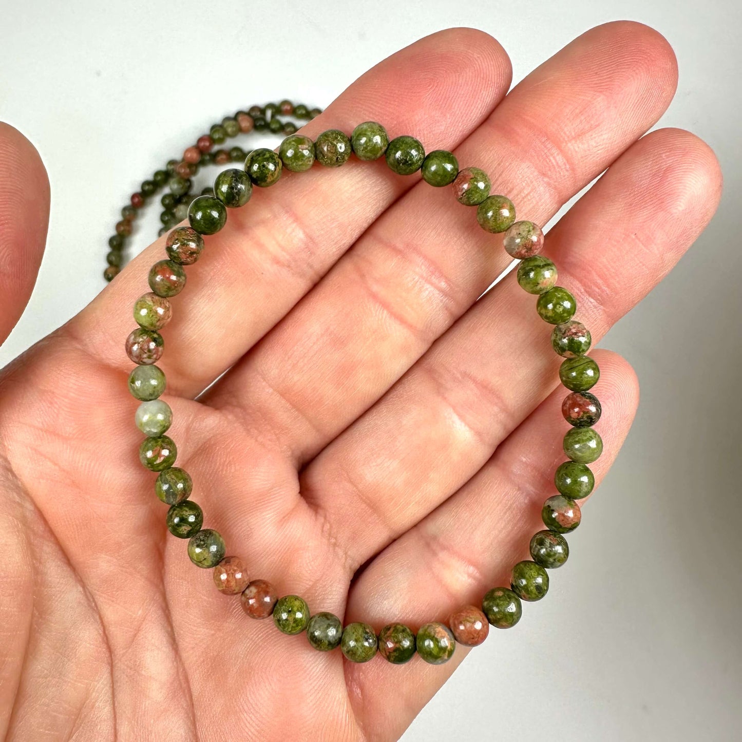 En hånd holder et unakit armbånd med grønne og lyserøde perler på en hvid baggrund.