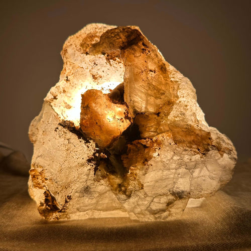 Klump af mineral med indvendig belysning, der fremhæver de varme og gyldne nuancer, placeret på en tekstureret overflade.