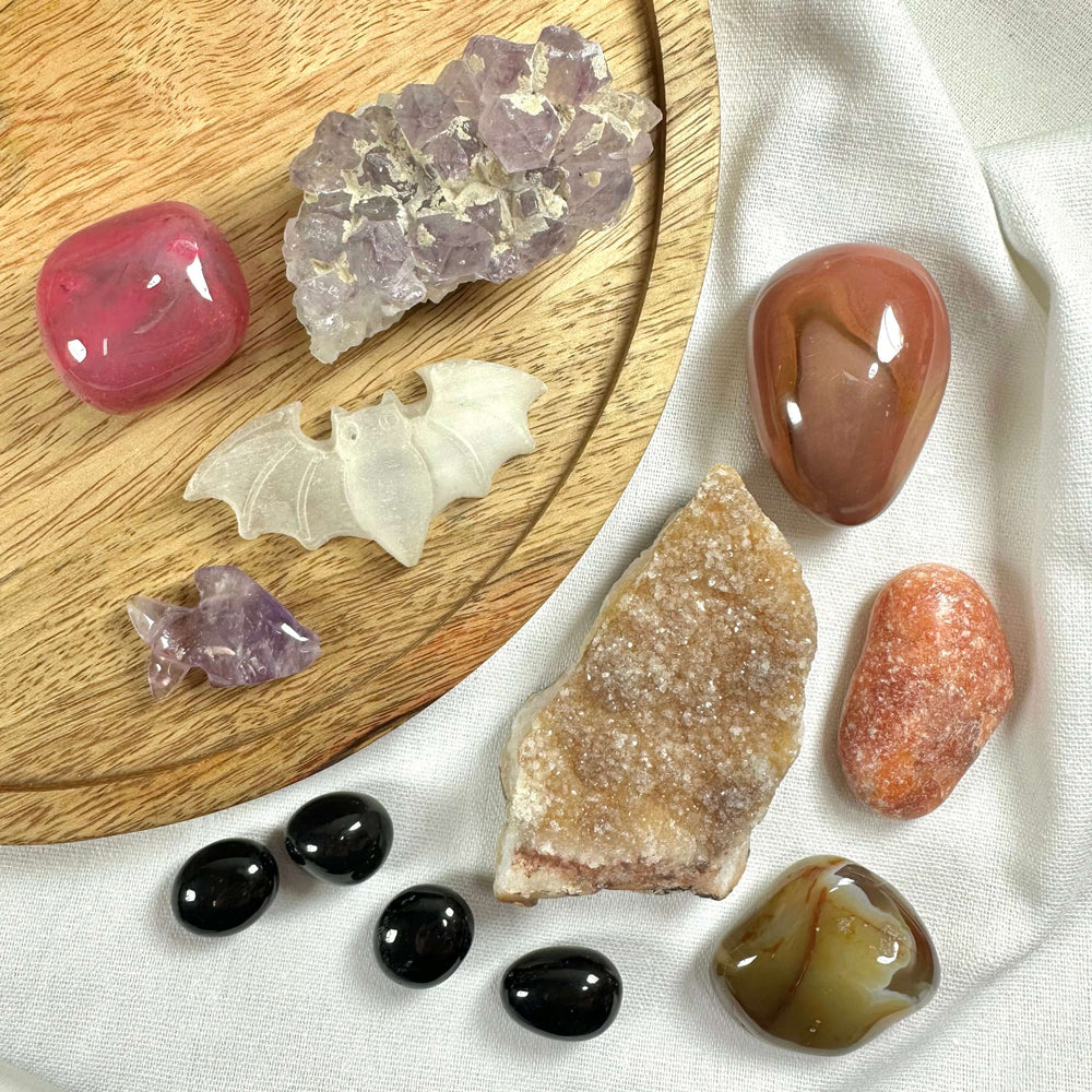 Et stemningsfuldt billede af Black Friday kurv #5 – Halloween lot #5 – hvor ni forskellige krystaller og sten er smukt arrangeret på en træbakke og hvidt stof. Blandt stenene ses en udskåret flagermus i gennemsigtig krystal og en lille fisk i ametyst. Farverne spænder fra rød, orange og brun til sort og lilla, hvilket skaber en mystisk og æstetisk helhed.