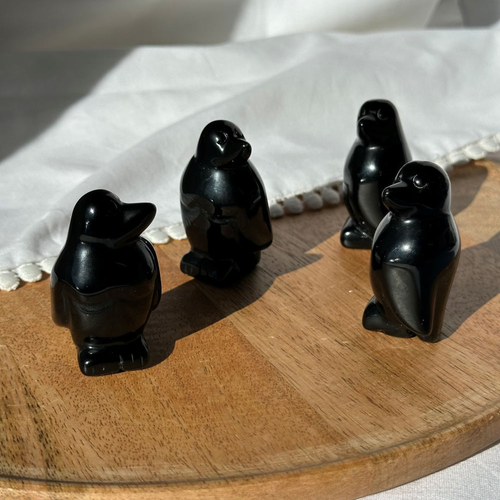 Fire små obsidian pingvin figurer står på en rund træplade, belyst af sollys.