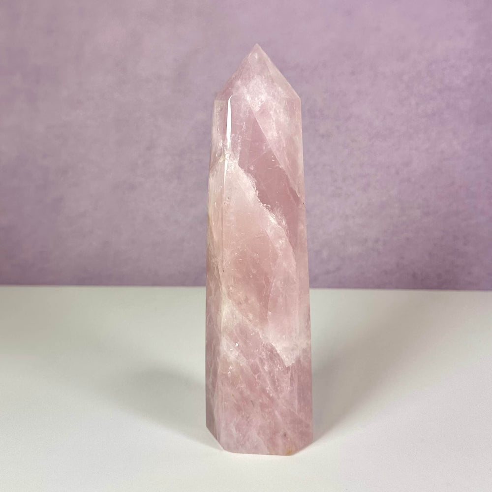 Et smukt rosenkvarts tårn står oprejst mod en lys lilla baggrund og en hvid overflade. Den lyserøde krystal er poleret med en glat, skinnende overflade og spidser til i en symmetrisk pyramideform. Kendt som kærlighedens sten symboliserer den selvomsorg, tilgivelse og følelsesmæssig heling. Et dekorativt og energisk redskab til meditation og harmoni.