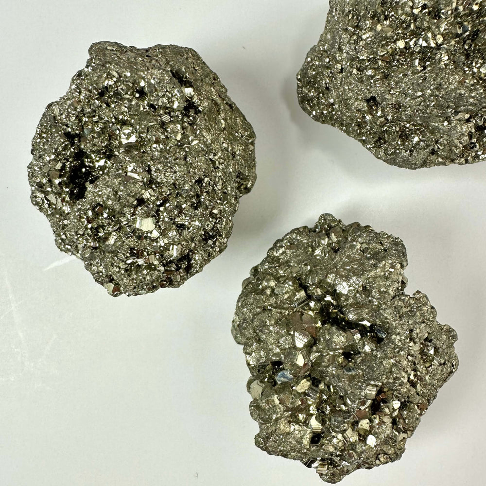 Tre skinnende pyrit klynger ligger på en hvid baggrund. De messinggule mineraler glitrer med deres karakteristiske metalliske glans og komplekse krystalstrukturer. Hver klynge består af utallige små krystaller, som fanger lyset og fremhæver deres guld-lignende udseende. Pyrit, også kaldet narreguld, er både et populært samlerobjekt og et spirituelt redskab med forbindelse til solar plexus chakraet.