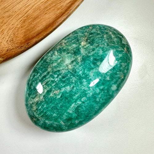 Amazonit palmstone afbildet i nærbillede på en lys baggrund, viser sin karakteristiske blågrønne farve med naturlige, marmorerede mønstre. Den glatte, ovale overflade reflekterer lyset, hvilket fremhæver stenens polerede finish. Bruges ofte i krystalhealing for sine beroligende og balancerende egenskaber, og siges at styrke kommunikation, intuition og forbinde hjerte og hjerne.