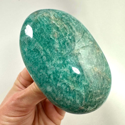 En hånd holder en glat, ovalformet amazonit palmstone med en smuk grøn-blå overflade. Stenen har naturlige mønstre og farvevariationer, som giver den et unikt og roligt udtryk. Amazonit er kendt for sine balancerende og beroligende egenskaber og anvendes ofte i krystalhealing og meditation. Den siges at fremme kommunikation, selvtillid og åbne hjertechakraet.