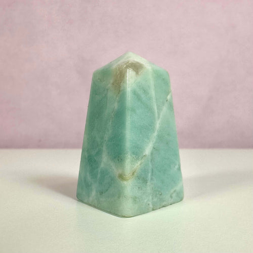 Amazonit obelisk i blågrønne og turkise nuancer står mod en blød rosa baggrund. Den polerede krystal er formet som en obelisk og symboliserer balance, klarhed og kommunikation. Den bruges til meditation, chakra-healing og spirituelle praksisser. Farvespillet i stenen skyldes naturlige mineraler og gør den både æstetisk tiltalende og energisk kraftfuld. Perfekt som dekorativ og spirituel genstand.