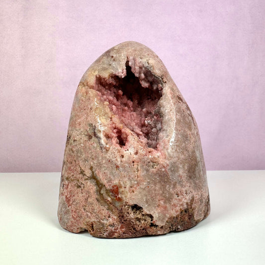 Pink Ametyst, unika – en imponerende rå krystal med naturlig geodeform og bløde pink og ferskenfarvede nuancer. Overfladen er glat og let skinnende, mens midten afslører funklende krystaller. Et sjældent stykke fra Patagonien, der kombinerer æstetisk skønhed med spirituel dybde og siges at fremme kærlighed, ro og følelsesmæssig healing. Fotograferet mod en rosa baggrund.