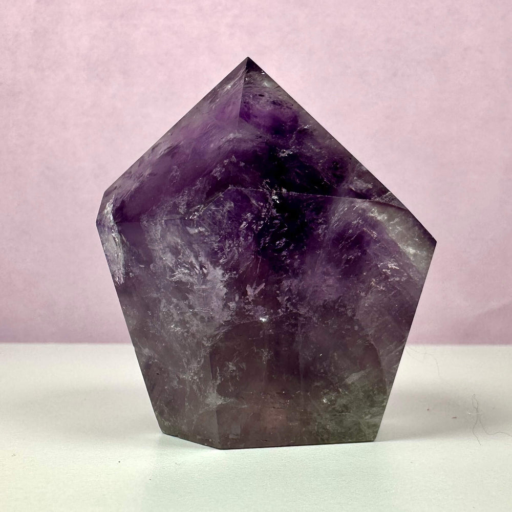 Ametyst spids med dybe violette nuancer, der elegant stråler mod en blød, lyserød baggrund. Den polerede krystal danner en tårnform med naturlige inklusioner, som giver den karakter og dybde. Ametysten symboliserer spirituel indsigt, renhed og beskyttelse samt skaber følelsesmæssig balance. Bruges ofte til meditation, energiarbejde og indretning som en dekorativ og beroligende energikilde.