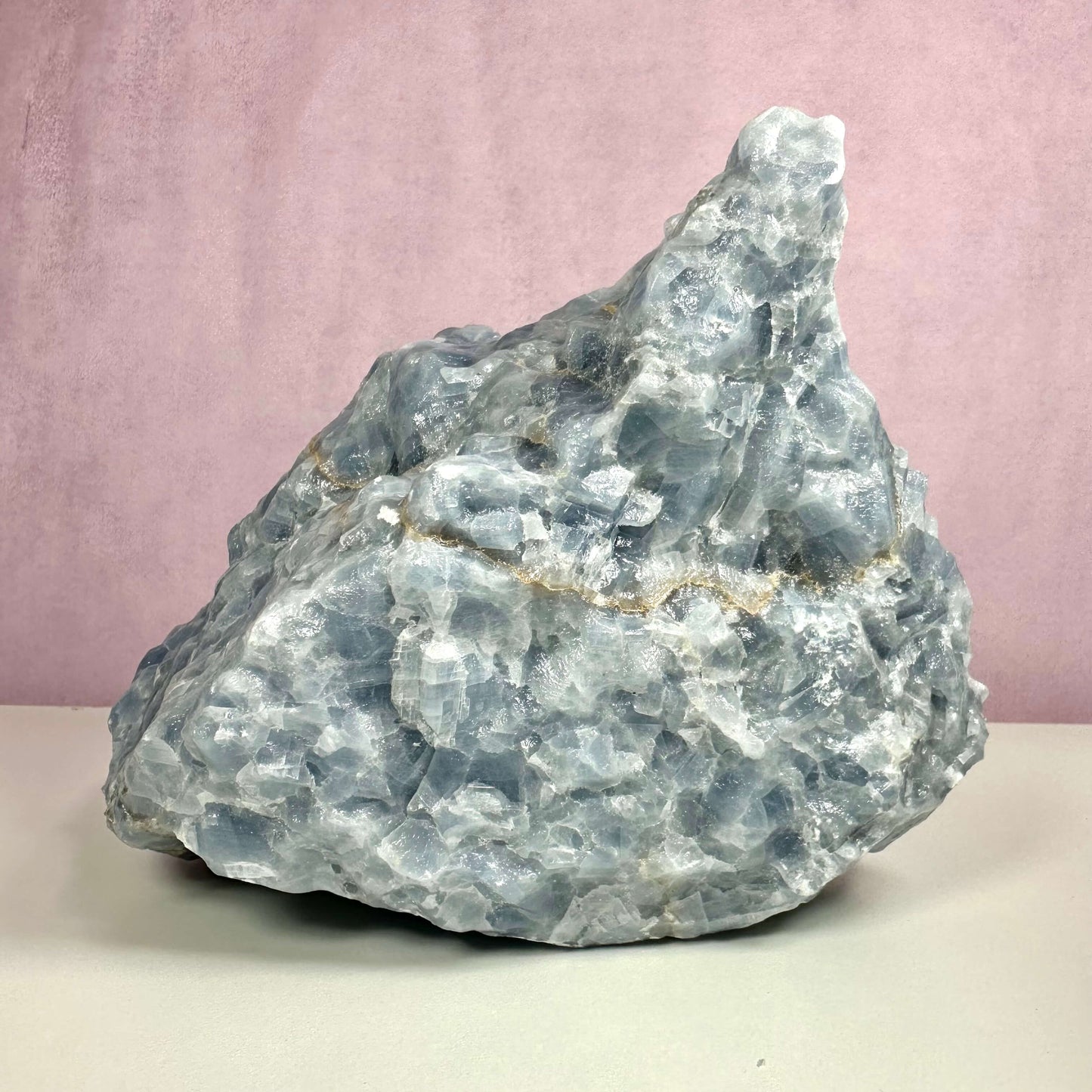 Rå blå Calcit nr 3 vises som en stor, ubehandlet krystal med ujævn overflade og lys blå farvetone. Den naturlige struktur afslører lag og glitrende partier, som fremhæver stenens unikke dannelse. Krystallen står placeret på en lys overflade med en blød rosa baggrund og formidler en følelse af ro, klarhed og æstetisk skønhed.