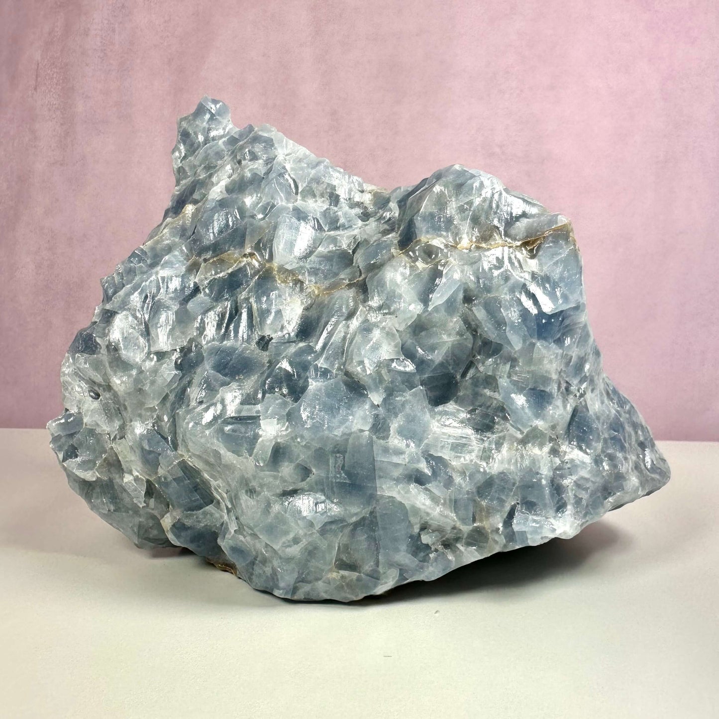 Rå blå Calcit nr 3 er en stor, naturlig krystal med en smuk lyseblå farve og ujævn struktur. Krystallen har en beroligende og balancerende energi og er særligt velegnet til meditation og spirituelt arbejde. Den forbindes ofte med halschakraet og styrker klar kommunikation. Baggrunden er rosa, hvilket fremhæver stenens kølige blå nuancer og dybde.