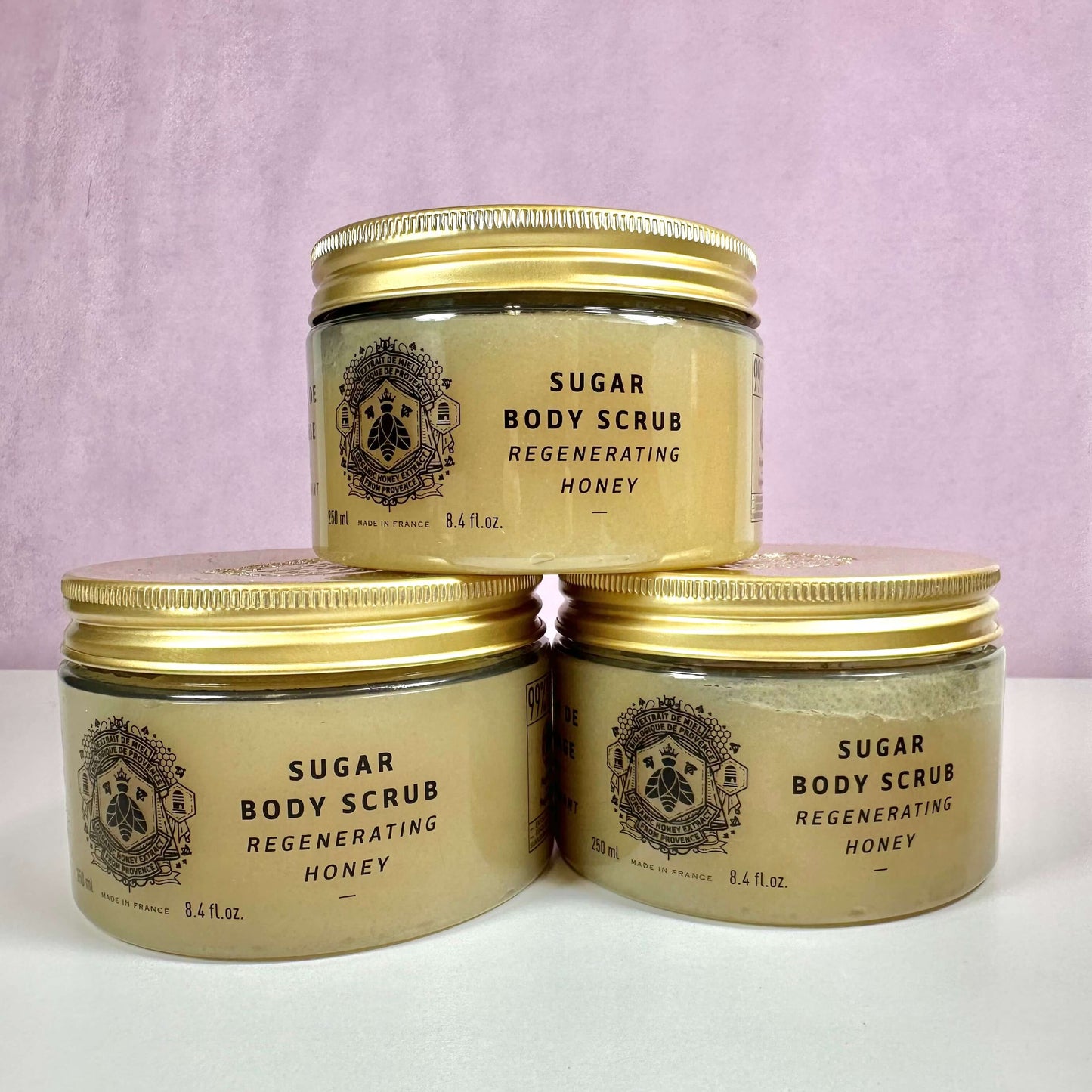 Tre krukker af Panier des Sens Sugar Body Scrub – Regenerating Honey er stillet op mod en rosa baggrund. Produkterne har gyldne låg og elegante etiketter med sort skrift og dekorativt emblem. Den sukkerskrub er designet til at regenerere huden og indeholder nærende honning. Hver krukke indeholder 250 ml og er fremstillet i Frankrig. Layoutet fremhæver produktets eksklusive og naturlige karakter.