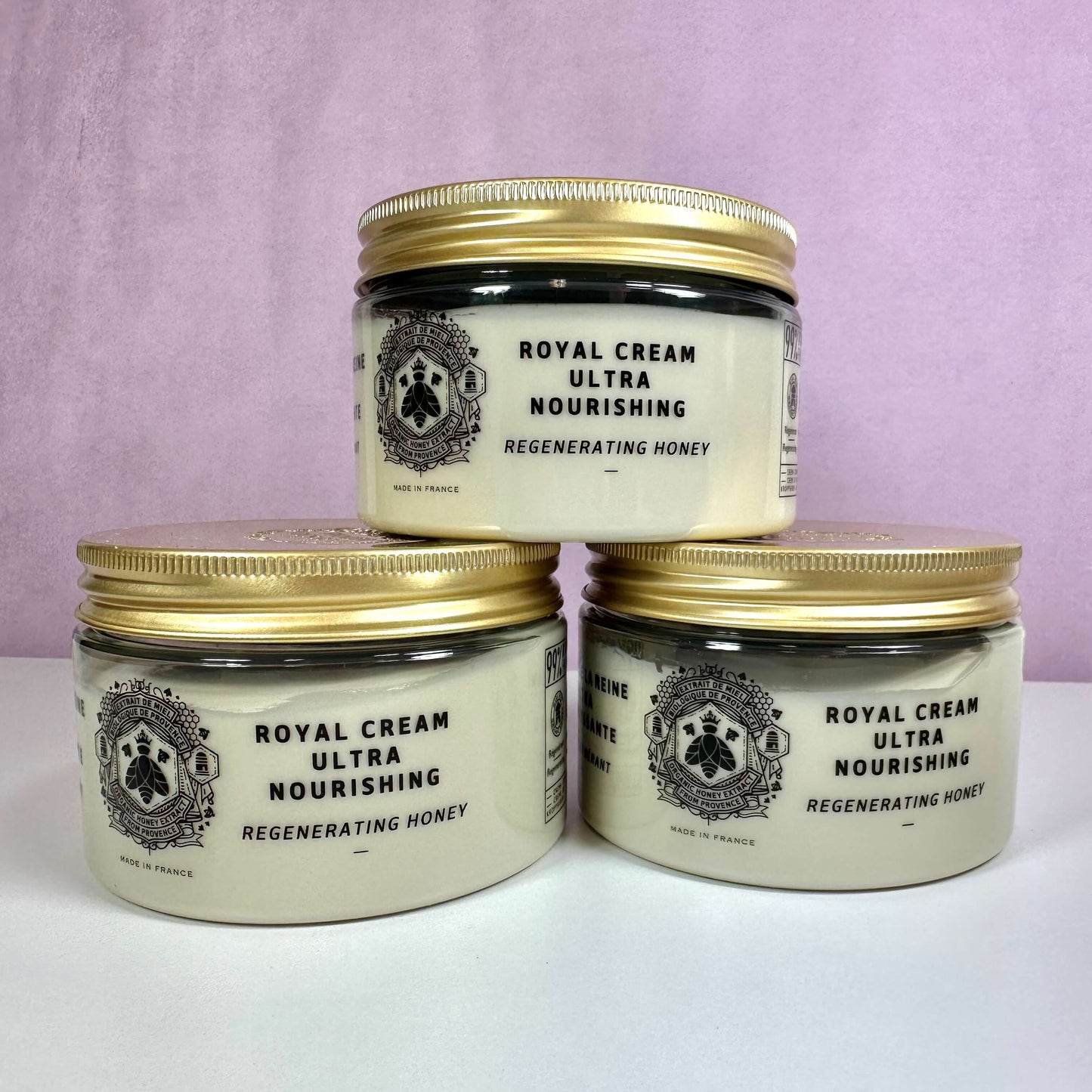 Tre krukker af Panier des Sens Royal Cream Ultra Nourishing er stillet op mod en lys lilla baggrund. Cremen er præsenteret i gennemsigtige plastbeholdere med gyldne låg og elegant sort skrift. Produktet indeholder regenererende honning og er lavet i Frankrig. Cremen er ideel til tør hud og indeholder økologisk honning, sheasmør og Royal Jelly, som giver intens fugt og næring.