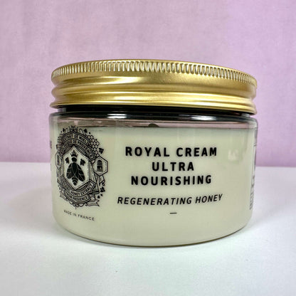 Et glas med Panier des Sens Royal Cream Ultra Nourishing står på en hvid overflade med en lyserød baggrund. Cremen kommer i en gennemsigtig beholder med gylden skruelåg og sort tekst, der angiver produktets navn og egenskaber. Designet er elegant med et fransk emblem foran og teksten "Made in France", som understreger produktets oprindelse.