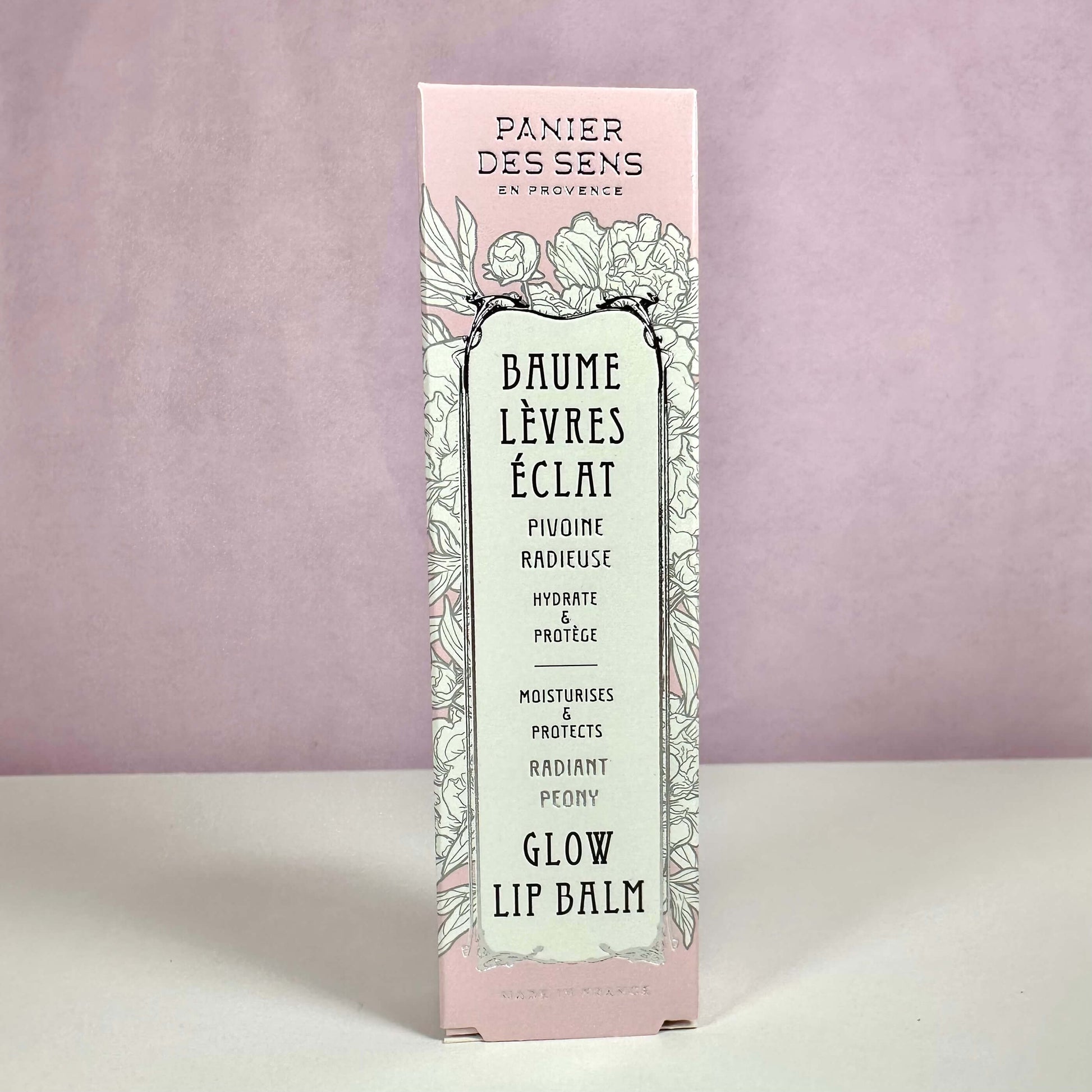 Produktet på billedet er Panier des Sens Glow Lip Balm, præsenteret i en elegant lyserød og mintgrøn æske med blomsterillustrationer. Teksten på æsken fremhæver, at læbepomaden fugter og beskytter læberne samt giver en naturlig glød. Designet understreger produktets franske oprindelse og botaniske inspiration. En læbepomade med pæonduft for bløde og strålende læber.