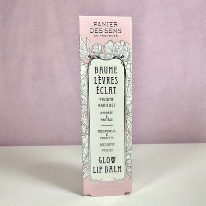 Produktet på billedet er Panier des Sens Glow Lip Balm, præsenteret i en elegant lyserød og mintgrøn æske med blomsterillustrationer. Teksten på æsken fremhæver, at læbepomaden fugter og beskytter læberne samt giver en naturlig glød. Designet understreger produktets franske oprindelse og botaniske inspiration. En læbepomade med pæonduft for bløde og strålende læber.
