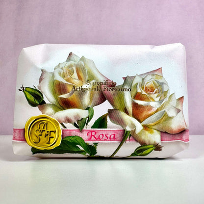 Fiorentino Rosa håndsæbe er elegant indpakket i papir med et smukt design af lyserøde og hvide roser. På forsiden ses teksten "Sapone Artigianale Fiorentino" samt et gult segl med initialerne "SAF". Den romantiske emballage udstråler italiensk luksus og klassisk charme, hvilket understreger sæbens eksklusive kvalitet. En duftende hyldest til naturlig skønhed og forkælelse af hænderne.
