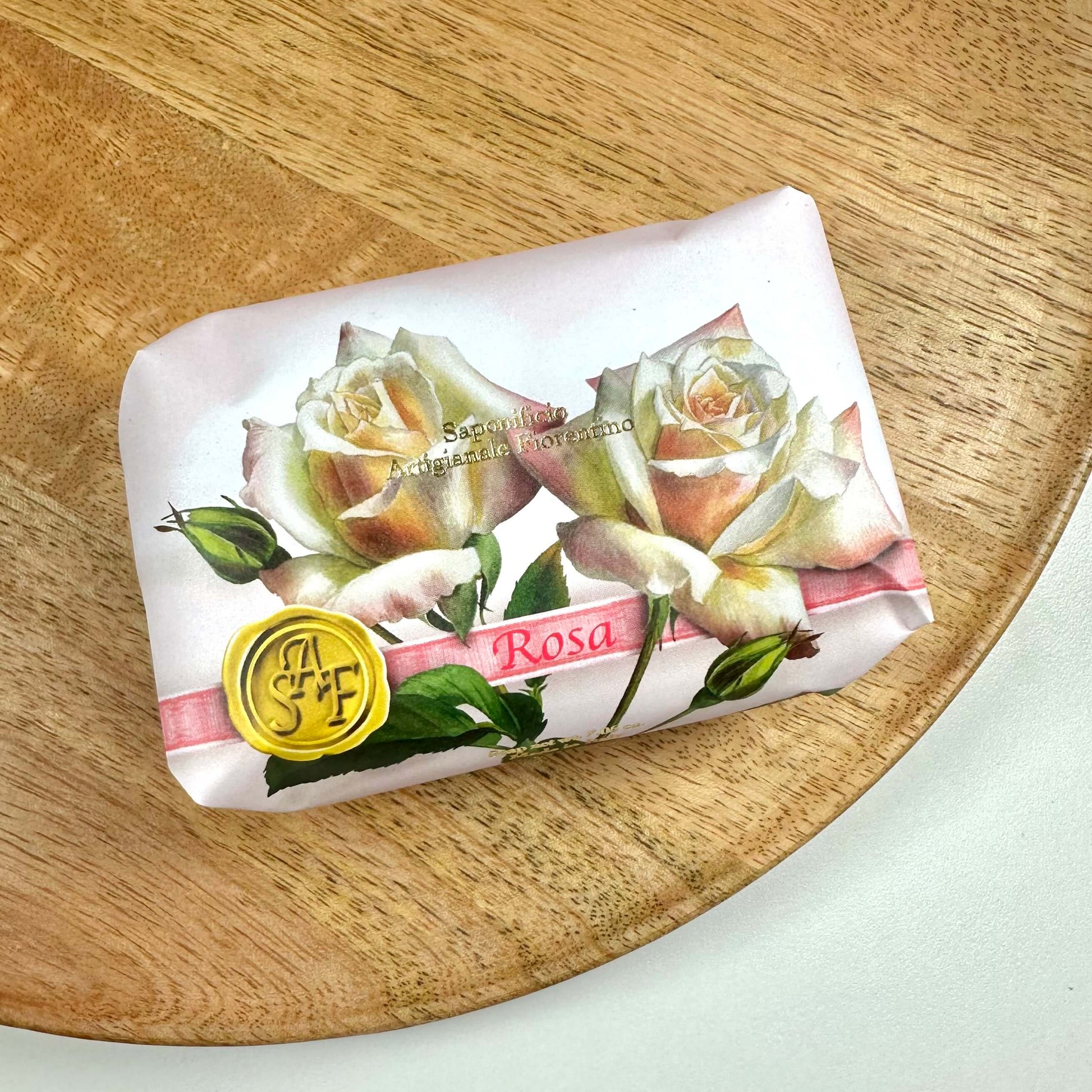 Fiorentino Rosa håndsæbe er smukt indpakket i en elegant lyserød emballage dekoreret med detaljerede illustrationer af roser og et gyldent segl med initialerne "S F". Sæben ligger på en rund træbakke og fremstår som en eksklusiv og romantisk gaveidé. Den bringer et strejf af italiensk luksus og naturlig skønhed ind i dine daglige plejerutiner.