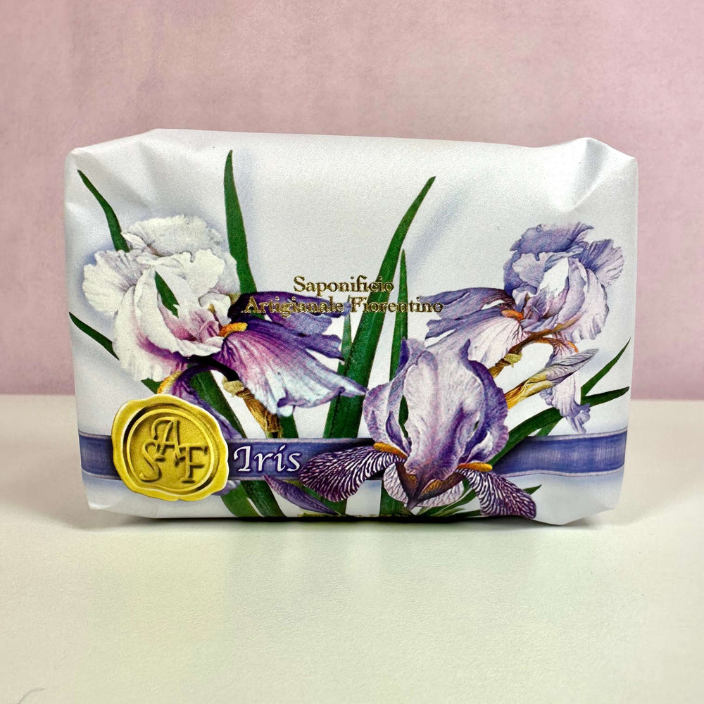 Fiorentino Iris Håndsæbe præsenteres i en elegant indpakning med smukke illustrationer af irisblomster i lilla og hvide nuancer. Produktet signalerer luksus og naturlighed med sin fine grafiske detaljering og subtil guldtekst. Den håndlavede vegetabilske sæbe er ideel til sensitiv hud og tilbyder både fugtgivende pleje og en sanselig duftoplevelse med inspiration fra traditionelt italiensk håndværk.