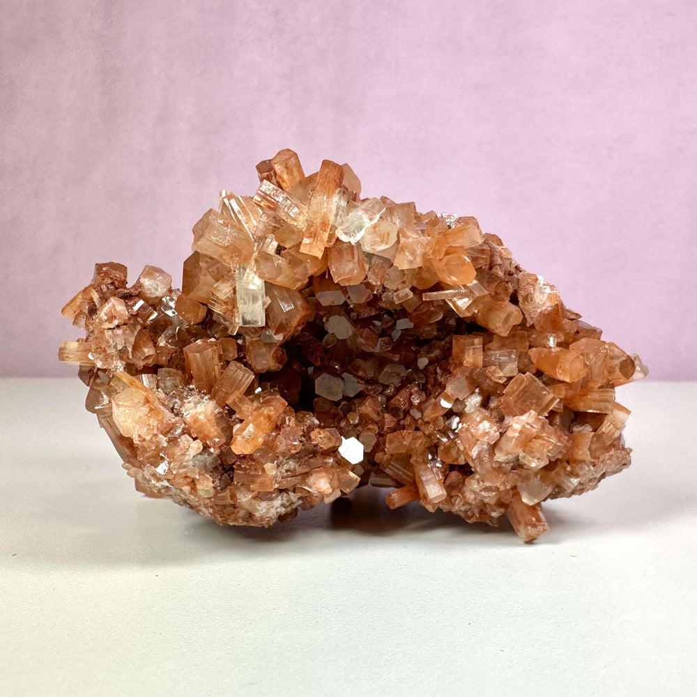 Aragonit klynge nr. 4 vises her med sin smukke, naturlige struktur af prismelignende krystaller i varme nuancer af brun, orange og hvid. Denne unikke mineralformation symboliserer jordforbindelse og indre balance. Den kan bruges til både spirituelt energiarbejde og som dekorativt element i hjemmet. Baggrunden er enkel og rosa, hvilket fremhæver klyngens naturlige skønhed.