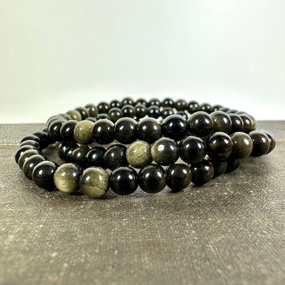 Gylden Obsidian armbånd med smukke, skinnende perler i mørke nuancer og gyldne reflekser, elegant arrangeret i et elastisk bånd. Armbåndet ligger på en rå overflade og fremhæver stenens naturlige glans. Kendt for sine healende egenskaber og stærke forbindelse til rodchakraet, tilbyder dette smykke både energetisk støtte og stilfuld beskyttelse i hverdagen.