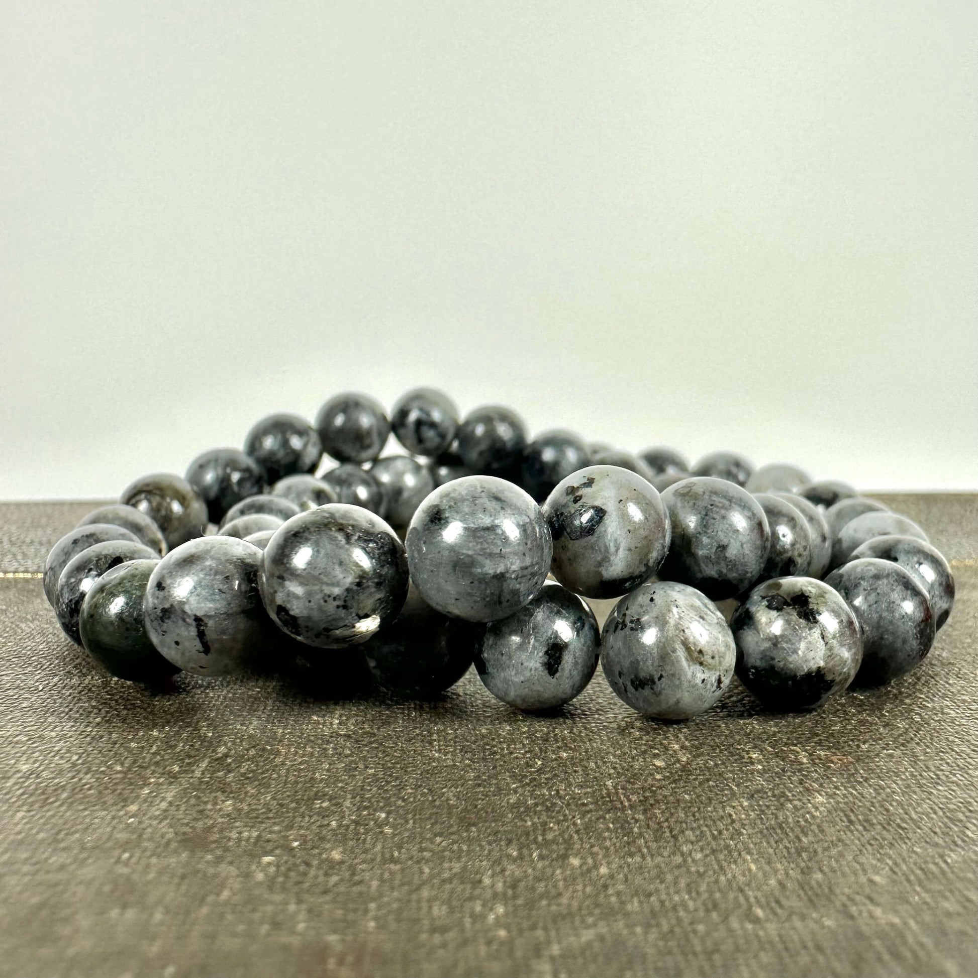 Et smukt Larvikit armbånd bestående af runde, polerede perler med mørkegrå og blålige nuancer, der reflekterer lyset med et subtilt skær. Armbåndet ligger elegant snoet på en rustik overflade og fremhæver stenens naturlige glans og struktur. Et ideelt smykke til dem, der søger både æstetisk skønhed og spirituel energi i deres hverdag.