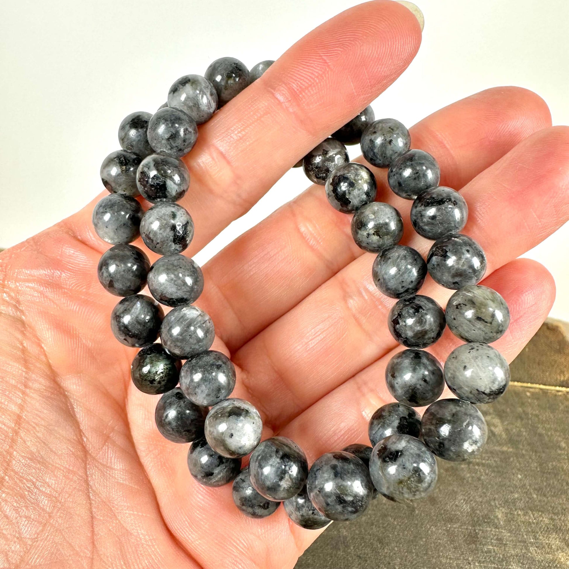 En hånd holder to smukke larvikit armbånd bestående af glatte, runde perler i nuancer af grå og sort med et let iriserende skær. Armbåndene udstråler en naturlig elegance og spirituel dybde. Larvikit siges at styrke intuition, beskytte mod negativ energi og skabe balance. Perfekt som både et stilfuldt smykke og et redskab til meditation og indre ro.