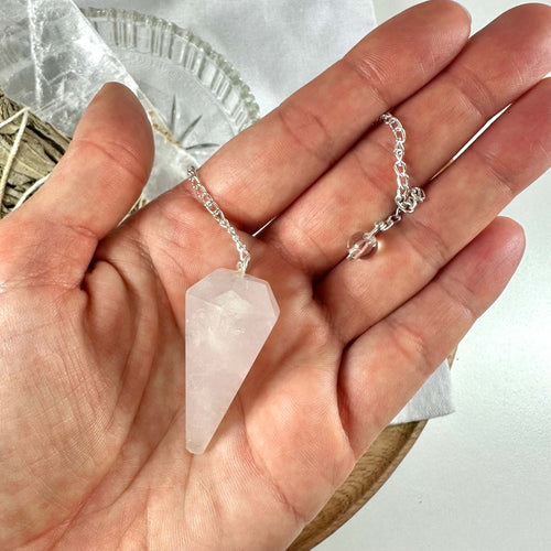 En hånd holder et rosenkvarts pendul med facetslebet, lyserød krystal og en sølvfarvet kæde. Pendulet symboliserer kærlighed, healing og følelsesmæssig balance og er kendt for sin forbindelse til hjertechakraet. Baggrunden inkluderer spirituelle elementer som hvidt stof og tørret salvie. Pendulet fremstår som et redskab til energiarbejde og spirituel praksis hos Krystalriget.dk.