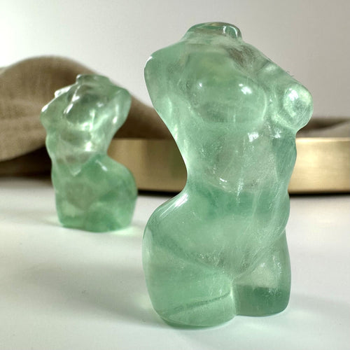 To grønne fluorit torsoskulpturer på en hvid overflade med en beigefarvet baggrund.