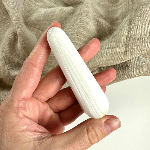 En hånd holder en glat, cylindrisk Scolesit stav mod en baggrund af beige stof.