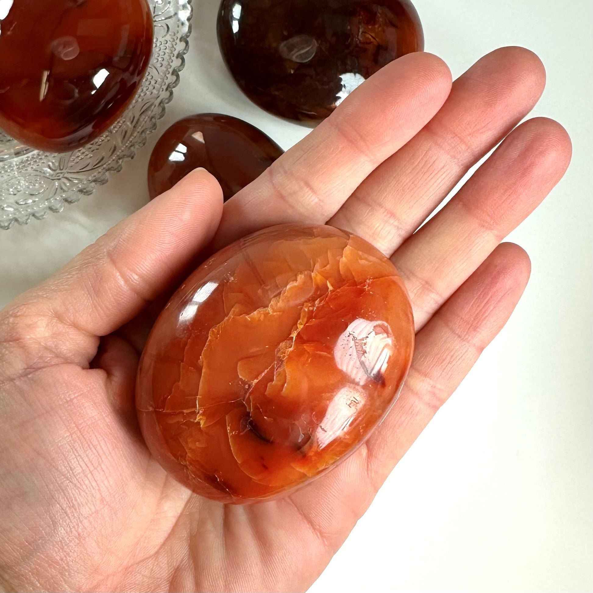 En hånd holder en glat, poleret karneol-palmstone med orange og røde nuancer. Flere sten vises i baggrunden.