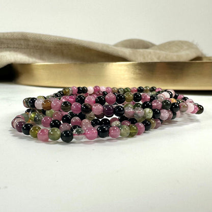 Et armbånd af flerfarvede perler ligger på en hvid overflade foran en gylden bakke med beige stof.