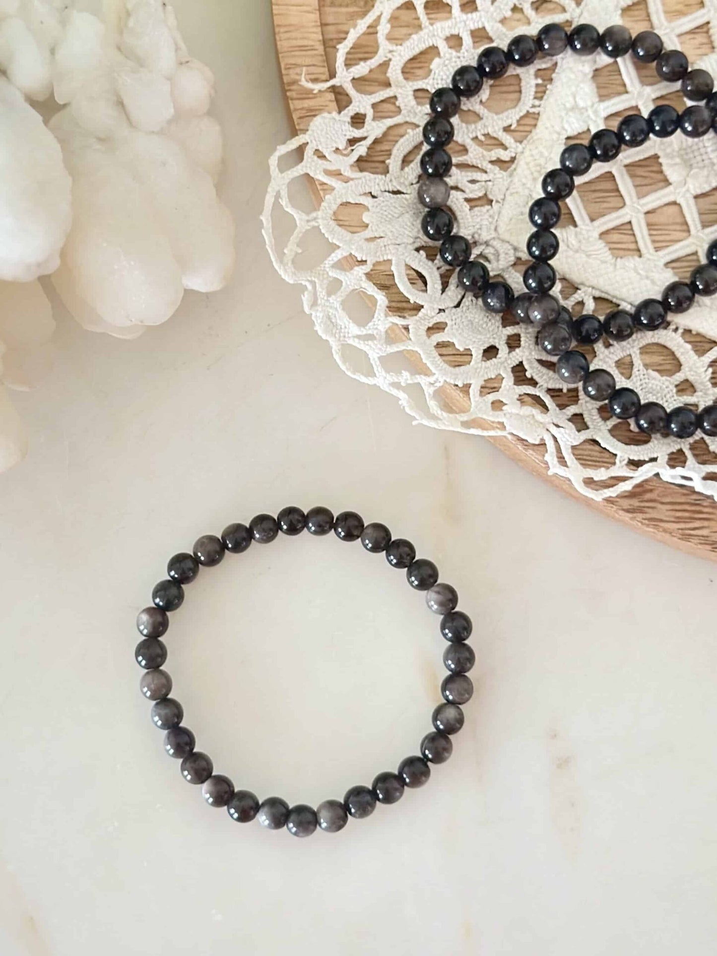 Obsidian sølv armbånd på en lysebrun flade med hvidt blondeunderlag.