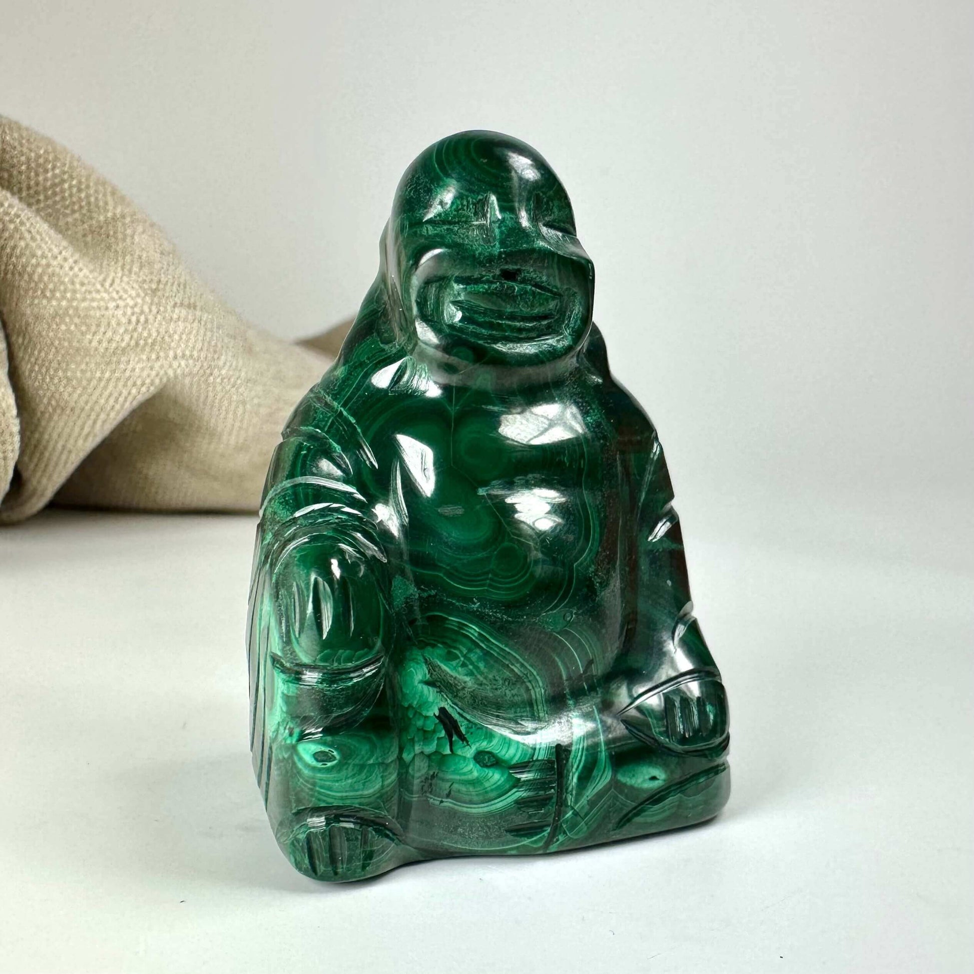 Malakit Buddha-statue med rund figur på hvid baggrund. Tæt på groft stofstykke i baggrunden.