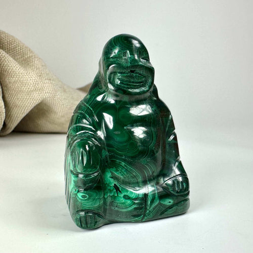 Malakit Buddha-statue med rund figur på hvid baggrund. Tæt på groft stofstykke i baggrunden.