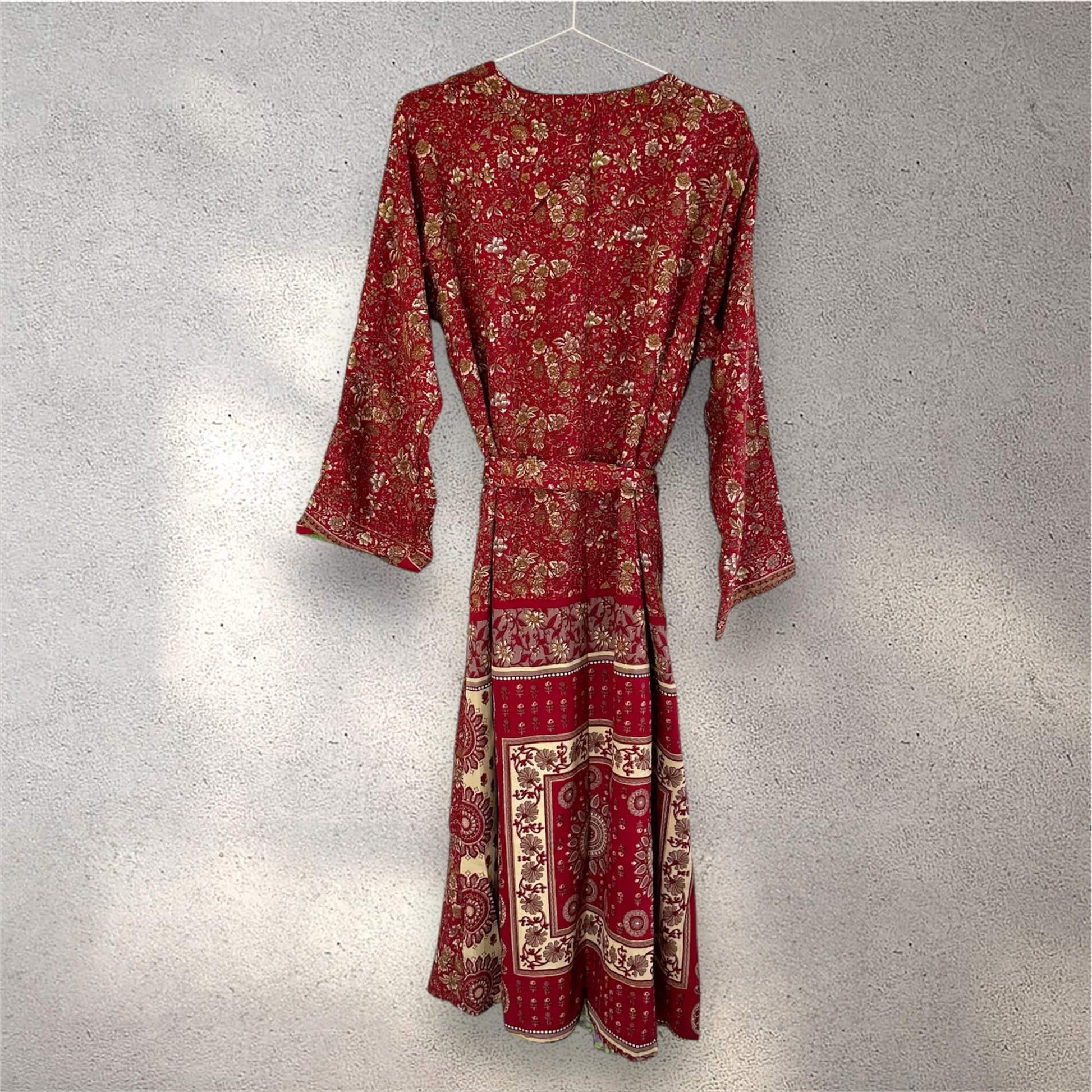 Rød kimono med blomster- og paisleymønstre hængende på en bøjle mod en grå væg.